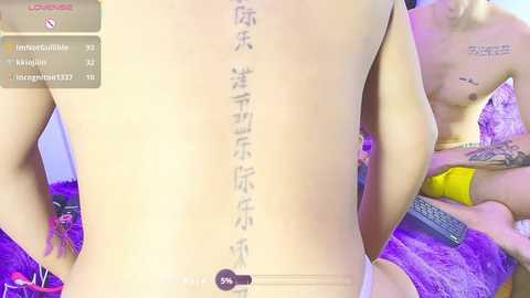 Video of a tattooed Asian man's back with Japanese characters, standing in a purple room. Another man with a tattoo on his chest sits on a bed, holding a remote.