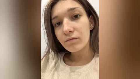 A close-up video of a young woman with light skin, dark hair, and a neutral expression, wearing a beige sweater, taken in a blurred, light-colored room.