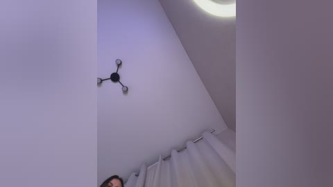 A video of a modern, minimalist bedroom corner with a white ceiling, a round light fixture, and a white curtain, featuring a person's partially visible head in the lower left corner.