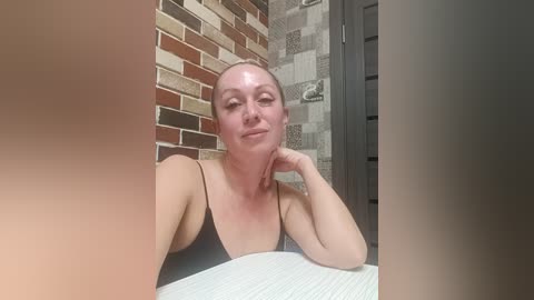 A video of a smiling, fair-skinned woman with a shaved head, wearing a black tank top, sitting in a bathroom with brick and tile walls.