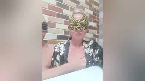 A video of a blonde woman in a leopard-print mask and floral robe, seated indoors with a brick wall background.
