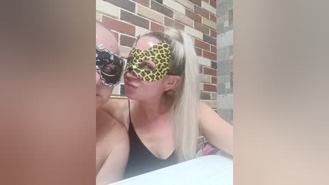 Video of a blonde woman with fair skin wearing a leopard-print masquerade mask, leaning against a brick wall with a white tiled floor.