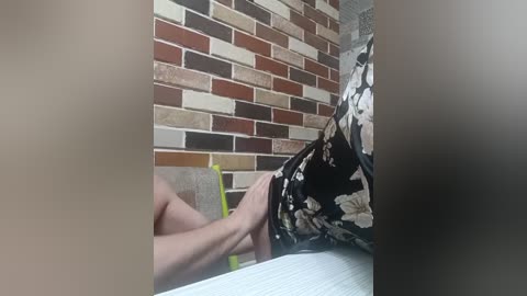 A video shows a person, likely male, seated at a table, wearing a floral-patterned shirt, with hands clasped in front. The background features a brick wall and a partially visible chair.