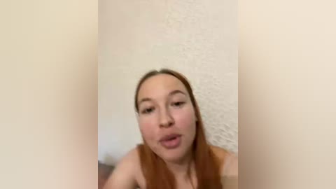 A blurry video of an Asian woman with light skin and straight, shoulder-length brown hair. She has a neutral expression, wearing a sleeveless top, against a textured off-white wall background.