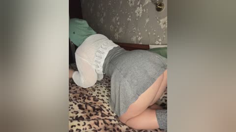 Video of a person in a leopard print blanket, wearing a gray skirt and a light blue sweater, lying on a bed with floral wallpaper in the background.