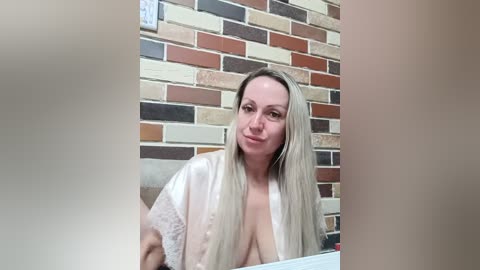 A video of a middle-aged woman with fair skin and long, straight blonde hair, wearing a white robe with lace trim, standing in front of a brick wall.