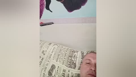 Video of a man with short hair lying in a bathtub, partially covered by a newspaper. The tub's white walls feature a colorful, abstract painting of a purple flower above.