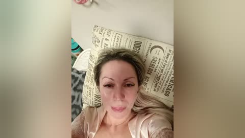 A video shows a smiling, light-skinned woman with long blonde hair lying on a bed covered with old newspaper. She wears a silky pink shirt and is surrounded by leopard print pillows.