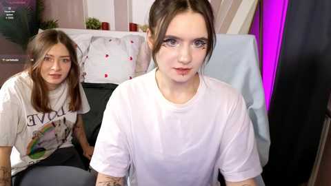 Video of two young women, one with brown hair and tattoos, wearing white T-shirts, seated indoors; background shows a card table with playing cards and a purple wall.