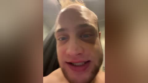 Video of a balding, smiling man with fair skin and a trimmed beard, partially obscured by out-of-focus objects in the foreground.