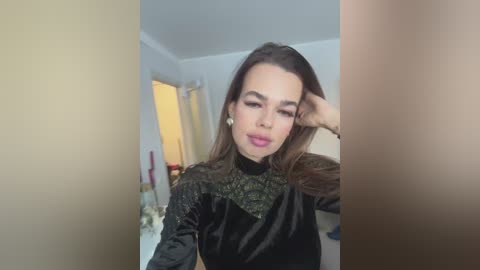 Video of a young woman with long brown hair, wearing a black velvet dress with gold accents, posing in a dimly lit room with a bed, curtains, and a doorway in the background.