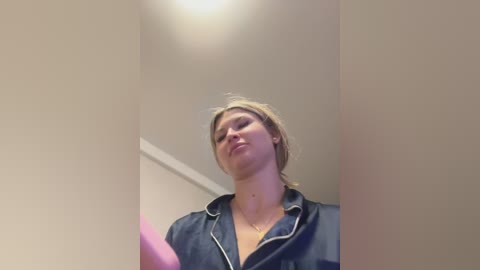 Video of a young Caucasian woman with fair skin and blonde hair, wearing a dark blue denim jacket with a zipper, standing indoors under a ceiling light.