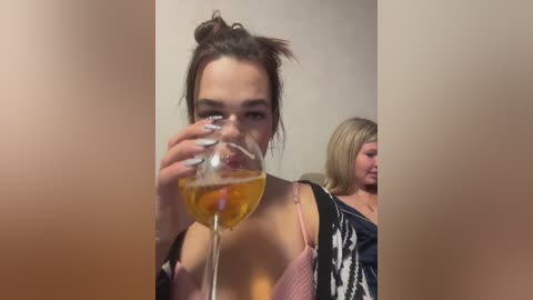 Video of a young woman with light skin and dark hair in a messy bun, wearing a pink bra, holding a glass of amber-colored drink, with another woman in the background.