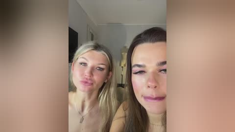 Video of two women kissing each other's lips, one blonde and one brunette, in a dimly lit, modern bedroom with beige walls and minimal decor.