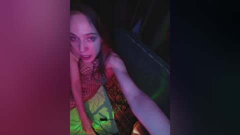 A video of a woman with fair skin and dark hair, wearing a red fishnet top and yellow pants, seated against a dark backdrop, lit with neon lights, creating a surreal, intimate atmosphere.