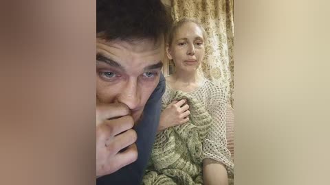 Video: A man with short brown hair, blue eyes, and a serious expression sits on a patterned couch, while a woman with blonde hair, fair skin, and a light beige crochet top sits next to him, looking concerned.