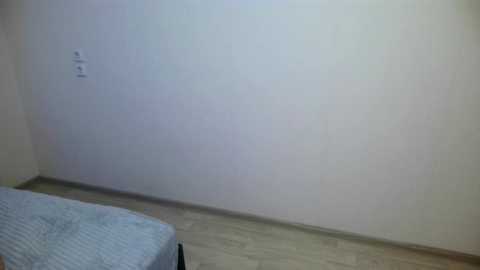 A video of a simple, sparsely furnished bedroom with a beige wall, a single bed with a striped blue blanket, and a light wooden floor.