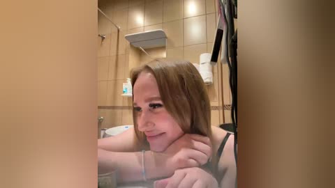 Video of a young woman with fair skin and brown hair, smiling, in a bathroom with beige tiles and a white sink.