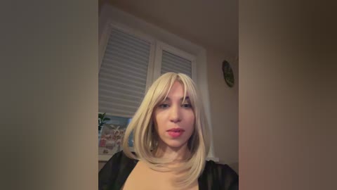 Video of a light-skinned, blonde woman with straight hair, wearing a black robe, standing in a beige room with white blinds and a potted plant on a windowsill.