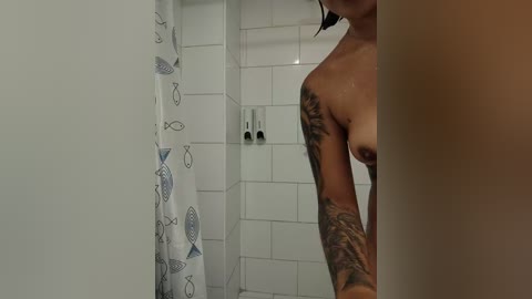 Video of a topless woman with wet, dark hair, standing in a white-tiled shower with a fish-patterned shower curtain. She has a large tattoo on her left arm.