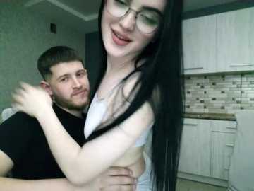 Video of a smiling, pale-skinned, black-haired woman with glasses, wearing a white tank top, hugging a bearded man in a black shirt in a modern kitchen with white cabinets and a tiled backsplash.