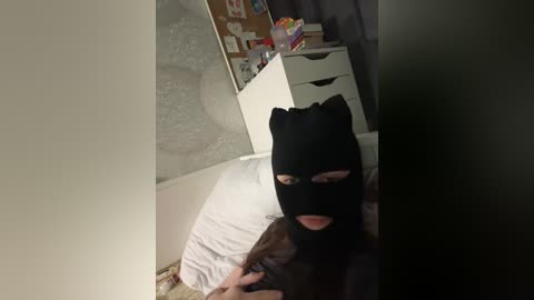 Video of a person wearing a black Batman mask, lying on a bed with white sheets, surrounded by a cluttered room with a white dresser and a corkboard.
