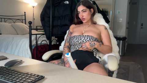 Video of a tattooed woman in a leopard-print strapless top, black shorts, and a white fur jacket, sitting at a desk in a modern bedroom.