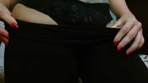 Video of a person's torso, hands pulling down black leggings, revealing lace underwear, red-painted nails, light skin tone, blurry background.