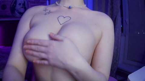 Video of a topless woman with pale skin and medium-sized breasts, covering them with her hands. She has a small heart tattoo on her chest and two star tattoos on her shoulder. Background includes a poster, curtains, and a door.