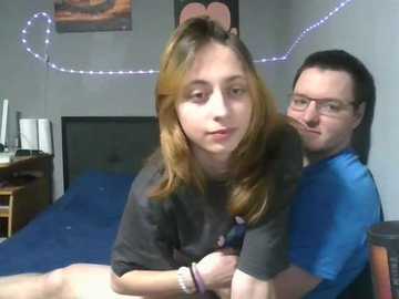 Video of a young woman with light brown hair in a dark shirt, leaning forward on a blue bed. A man with glasses and dark hair sits behind her, looking at the camera.