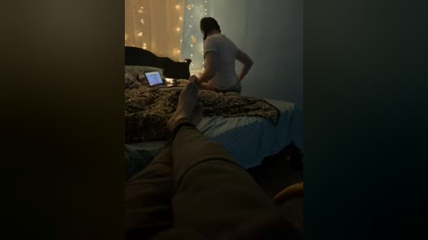 Video of a dimly lit bedroom showing a man in a white shirt working on a laptop, while a woman's legs are visible in the foreground.