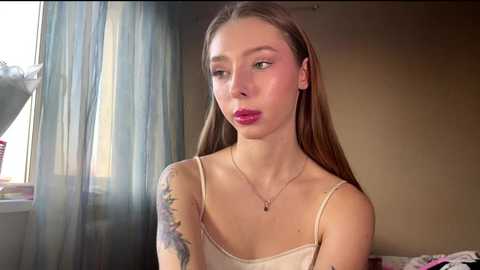 Video of a young woman with light skin, long brown hair, and pink lips, wearing a beige camisole, sitting in a room with blue curtains and a window.