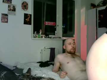 A video of a bald, fair-skinned, nude man with a beard lying on a bed, eyes closed, in a dimly lit, cluttered room with photos on the wall and a white radiator.