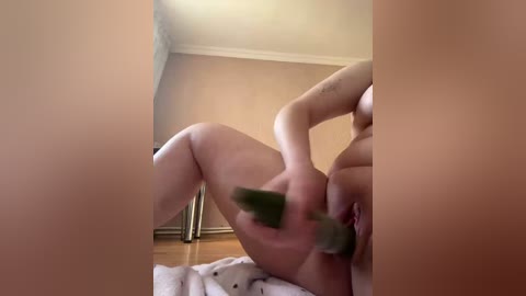 A video of a nude, light-skinned woman with a tattoo on her left arm, using a green dildo on herself in a dimly lit room with beige walls and wooden floors.