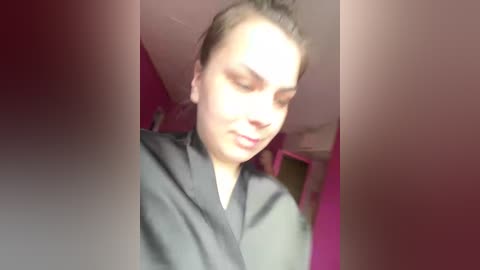 Video of a young woman with light skin and brown hair in a bun, wearing a black robe, sitting in a dimly lit room with red walls.