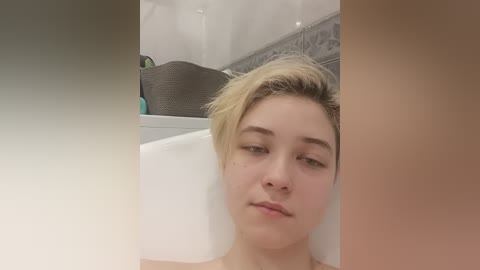 Video of a young Caucasian woman with short, blonde hair, sitting in a bathtub, looking relaxed. The background features a tiled wall and a gray towel hanging.