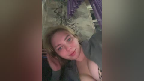 A video shows a young woman with light skin and shoulder-length blonde hair, wearing a leopard-print top. She's indoors, surrounded by purple curtains and a cluttered background.