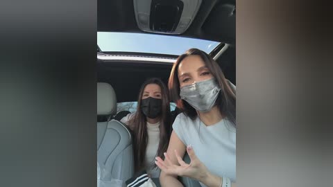 Video of two women wearing masks, sitting in a car. One has long dark hair, the other medium length. They appear relaxed and engaged in conversation.