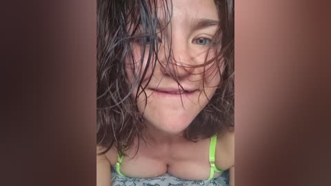 Video of a young woman with wet, dark hair covering her face, wearing a green bra with light gray lace. Her skin is pale, and she has a slight smile. The background is blurry, brownish.