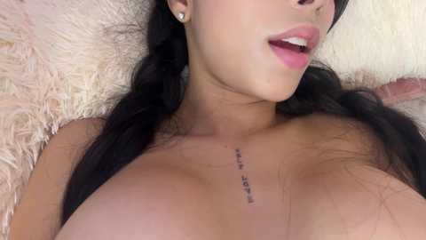 Video of a topless woman with long black hair, light skin, wearing small earrings, lying on a plush cream rug, mouth slightly open, showing a tattoo of text on her collarbone.