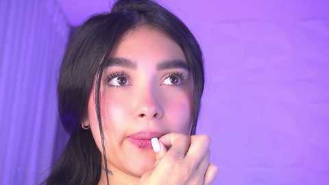 Video of a young woman with long black hair, light brown skin, and large eyes, applying lipstick in a soft purple-lit room, with a subtle white wall in the background.
