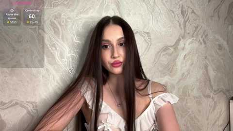 Video of a young woman with long brown hair, fair skin, and a slender physique, wearing a white off-shoulder top. She stands against a textured, light-colored marble wall.