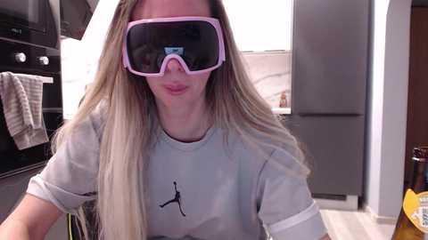 Video of a blonde woman wearing a pink virtual reality headset, grey T-shirt with a black silhouette, in a modern kitchen with dark cabinets and light countertops.