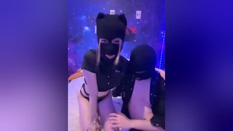 A video of two people in black cat masks, wearing black and white outfits, sitting on a bed with a starry blue backdrop.