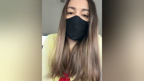 A video of a young woman with long, straight brown hair, wearing a black face mask, a yellow blouse, and a red tie. She has light skin and is standing indoors against a blurred background.