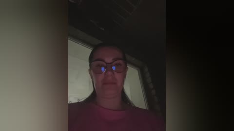 A dimly lit video of a woman with long, dark hair, wearing glasses and a red shirt, standing in front of a dark, blurry background.