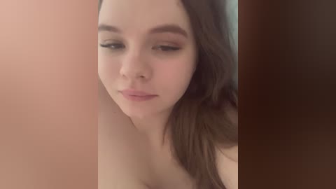 A close-up video of a young woman with fair skin, brown hair, and light makeup, partially obscured by a blurry, brownish background. She appears to be topless, with a neutral expression.