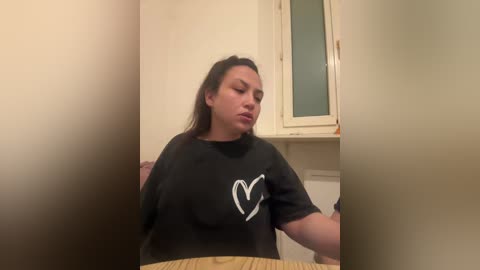 Video of a plus-size woman with medium skin tone, long brown hair, wearing a black t-shirt with a white heart graphic, sitting at a wooden table in a dimly lit room with beige walls and a frosted window.