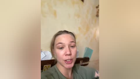 A video of a young woman with light skin and brown hair tied back, wearing a green shirt. She's indoors, with a textured yellow wall and a brown couch behind her.