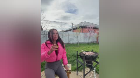 Video of a woman with long brown hair, wearing a bright pink jacket and gray pants, sitting on a patio, holding a glass, beside a charcoal grill, in a suburban backyard with a white fence and a red-brick house.